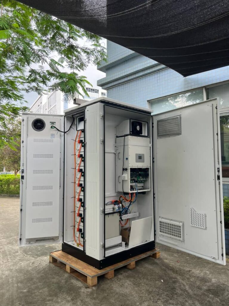 Open outdoor battery storage cabinet on wooden pallet showing internal wiring, cooling fan, and inverter equipment.